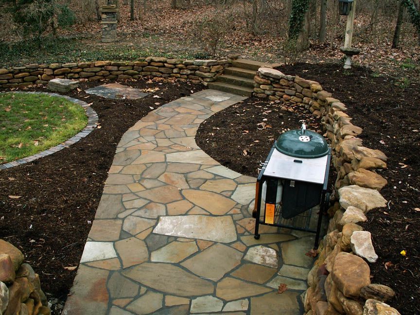 A stone path winds through a backyard, bordered by a low stone retaining wall and mulch, leading to small concrete stairs.