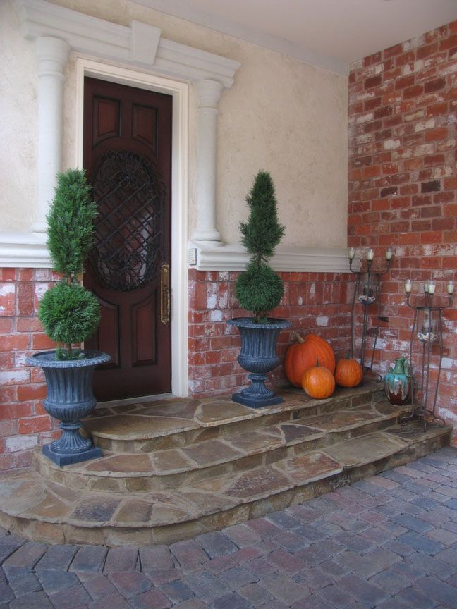 A wooden front door with a decorative oval window, flanked by two topiary plants in gray urns on stone steps with pumpkins.