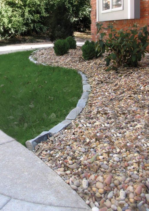 A stone border separates a green lawn from a garden bed filled with multicolored gravel and small shrubs by a brick wall.