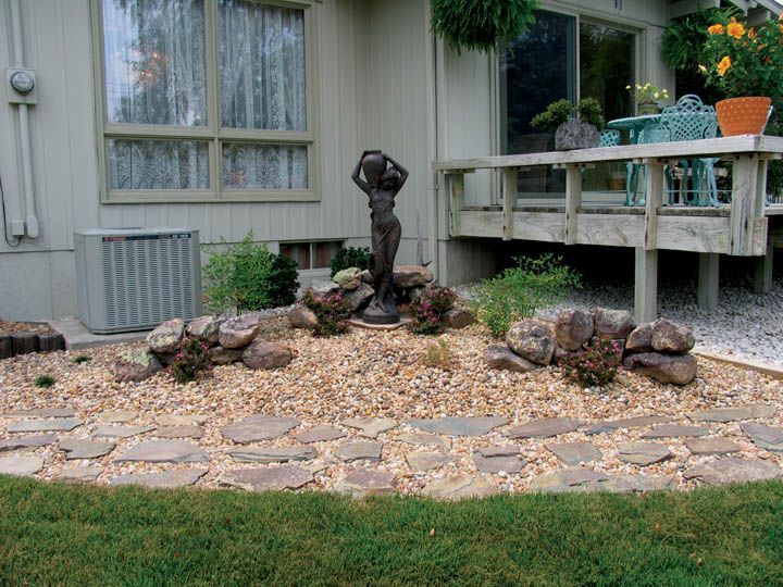 A landscaped rock garden bed with a central statue of a person, bordered by a stone path in front of a house.
