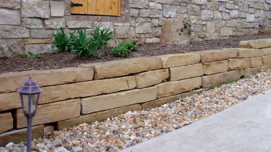 A stacked sandstone retaining wall borders a gravel pathway in front of a stone house with a wooden shutter.