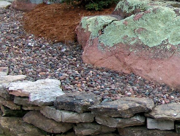 A stone retaining wall holds back a garden bed with dark gravel, pine needle mulch, and a large, lichen-covered boulder.