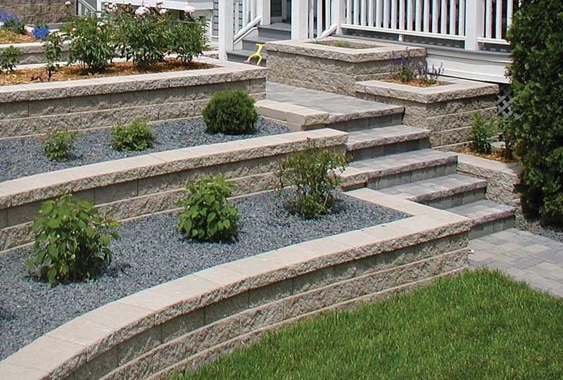 Tiered stone garden beds with shrubs, gravel, steps, and a white porch railing.