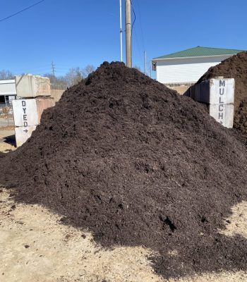 Large dark mulch pile outdoors with compost bins on either side and a blue sky background