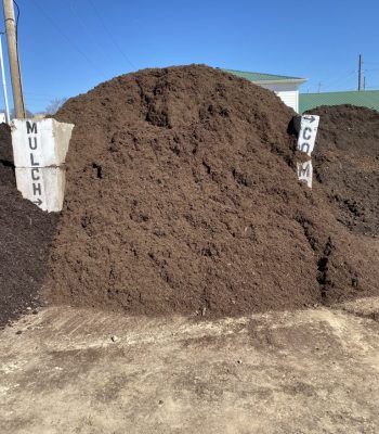Large pile of mulch at an outdoor yard, with white signs reading “MUCH” on the sides.