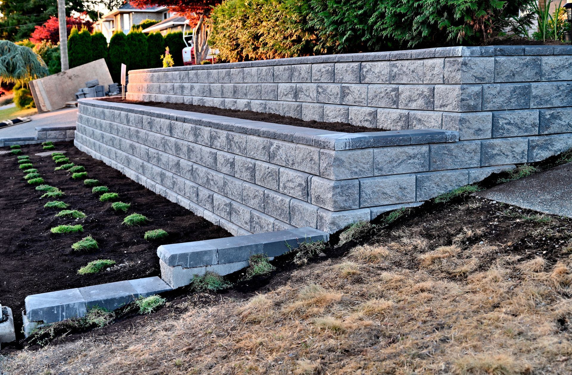 Gray block retaining wall beside a sloped landscaped yard with young plants and mulch