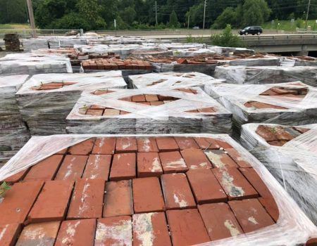 Stacks of red clay tiles wrapped in plastic on a roadside near trees and a parked car.