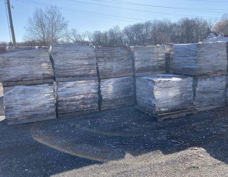 Stacks of wrapped pallets or bundles outdoors on a gravel lot under a clear sky.