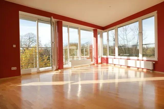 Empty room with glossy hardwood floors, large windows, and red walls, bathed in sunlight.