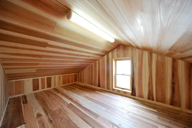 Wooden-paneled attic room with sloped ceiling, small window, and natural light.