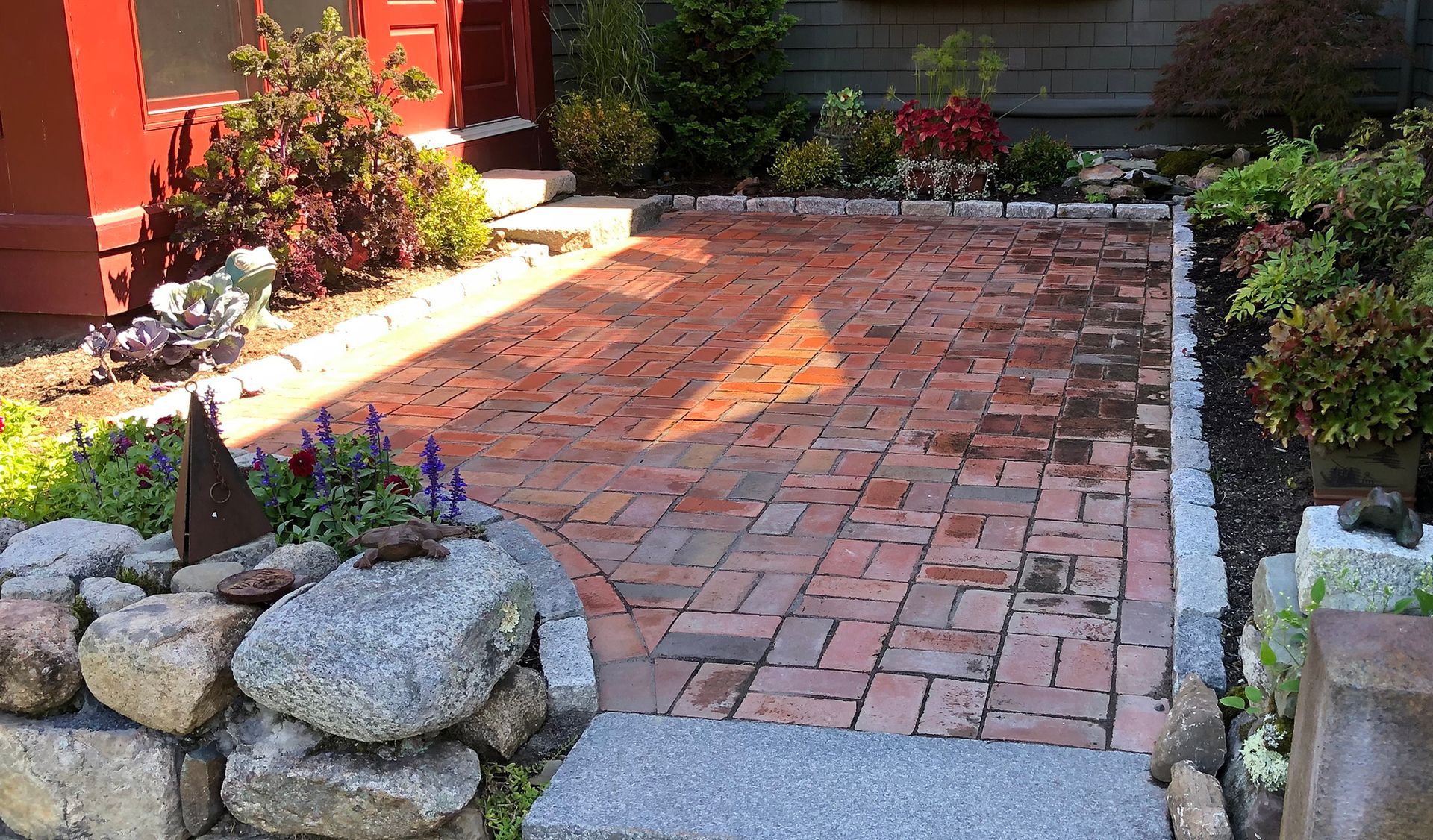 Two wooden benches sit on a circular red brick patio surrounded by lush green trees and shrubs.
