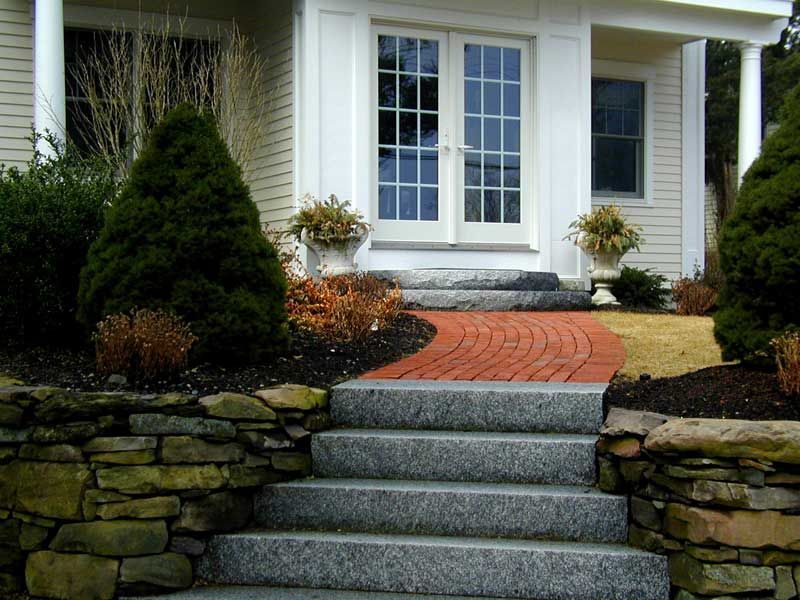 Stone steps lead up to a house entrance featuring a red brick walkway, white door, and side planters.