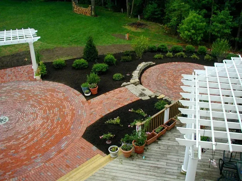 A high-angle view of two circular brick patios connected by a walkway in a garden with mulch beds and white pergolas.