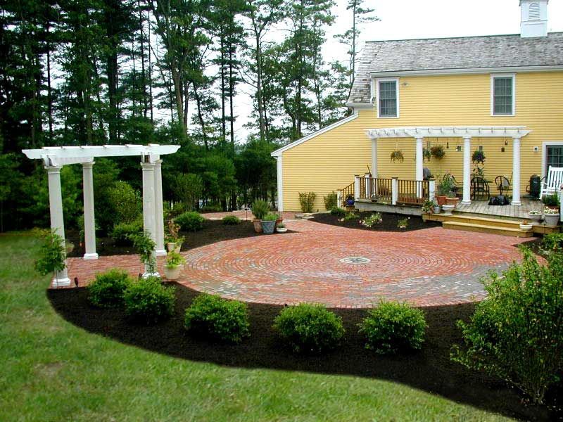 A circular brick patio with a white pergola in a backyard next to a yellow house with a deck and landscaping.