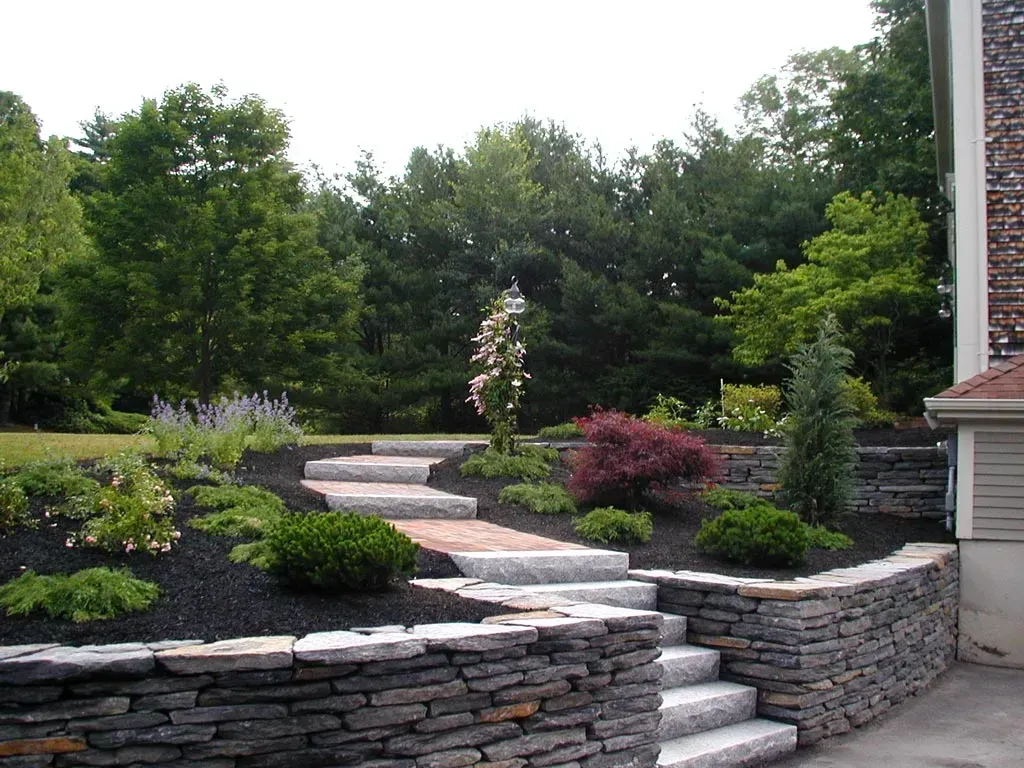 A stone retaining wall with matching steps leads up to a landscaped yard with trees, shrubs, and mulch.