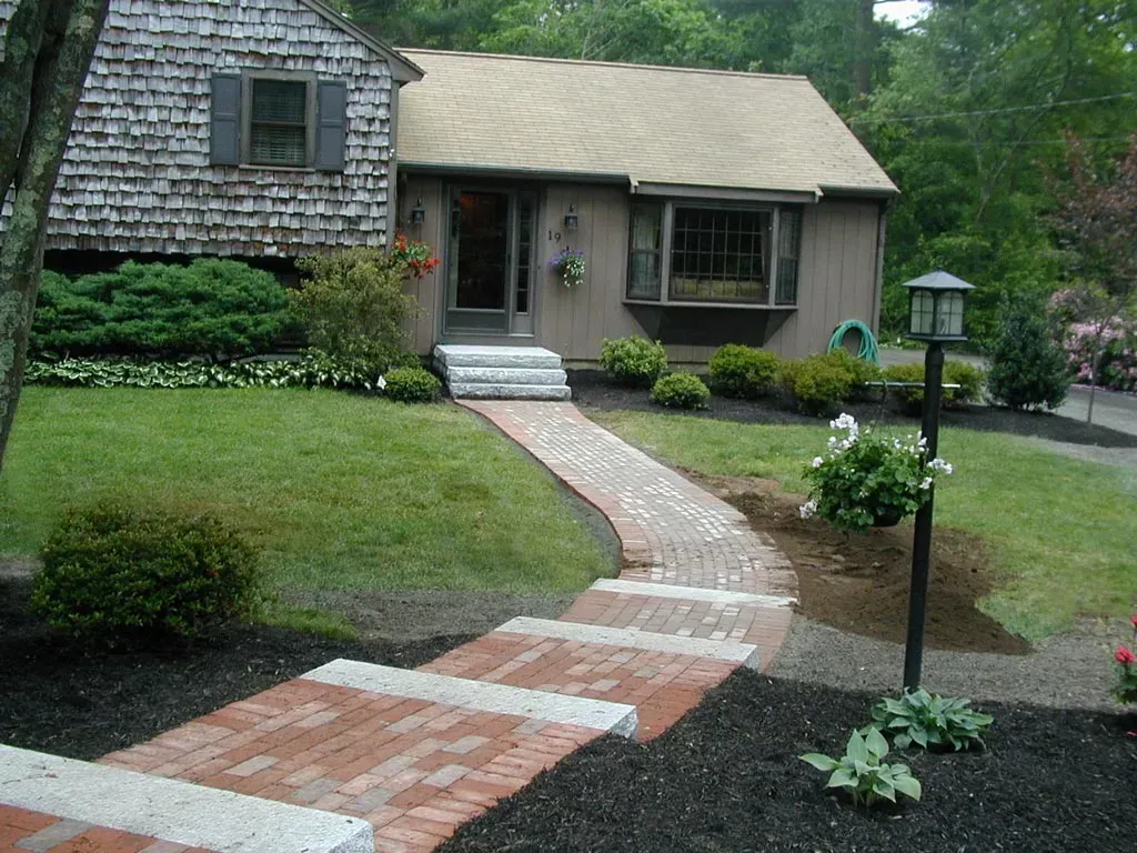 A brick walkway with stone accents leads to the front door of a suburban house with shingle siding and a bay window.