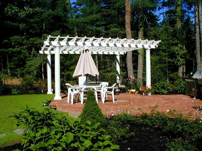 A white pergola with a dining set and umbrella on a red brick patio in a wooded backyard.