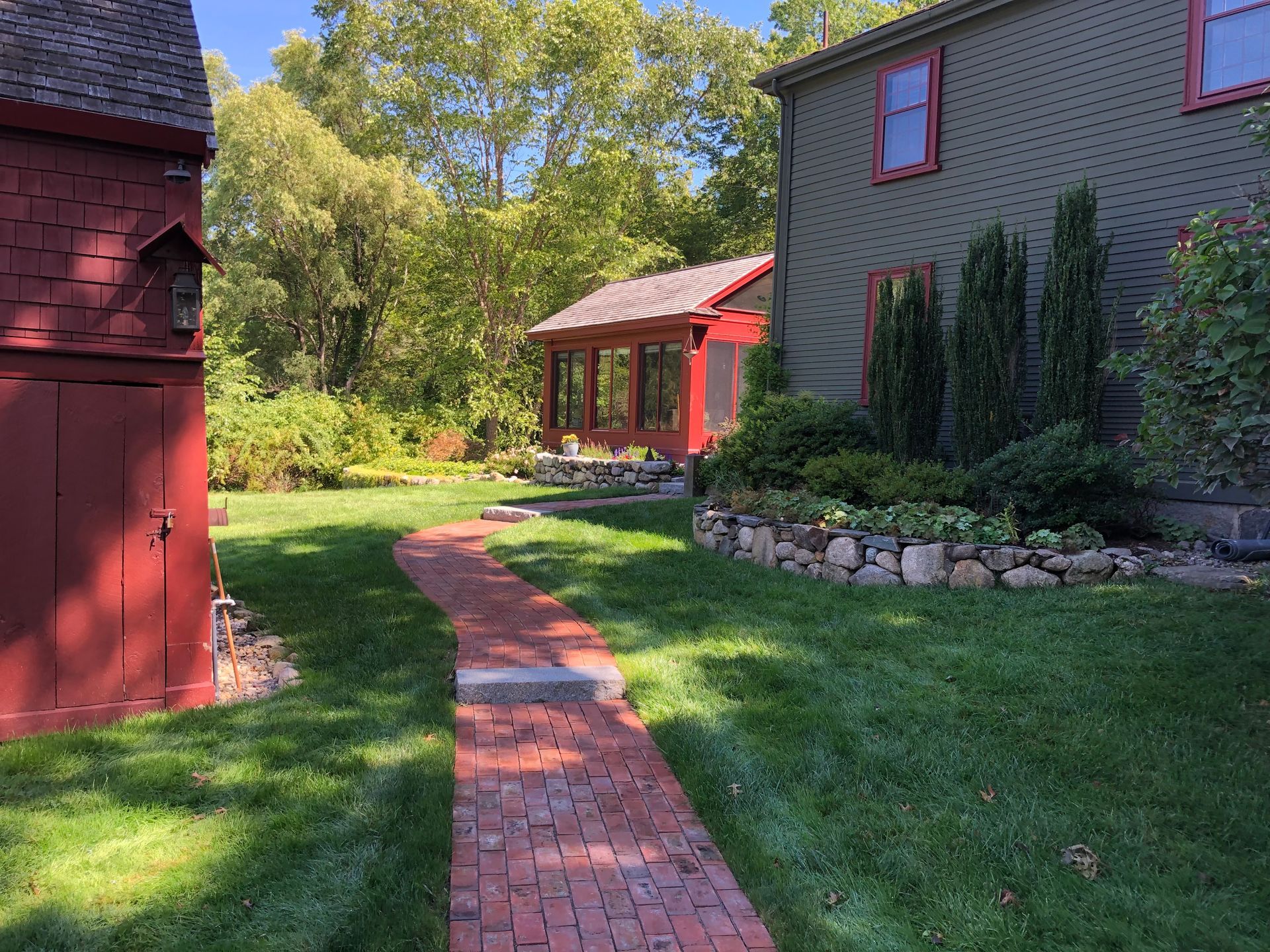 A brick path leads through a grassy yard between a red wooden outbuilding and a dark green house with a sunroom.