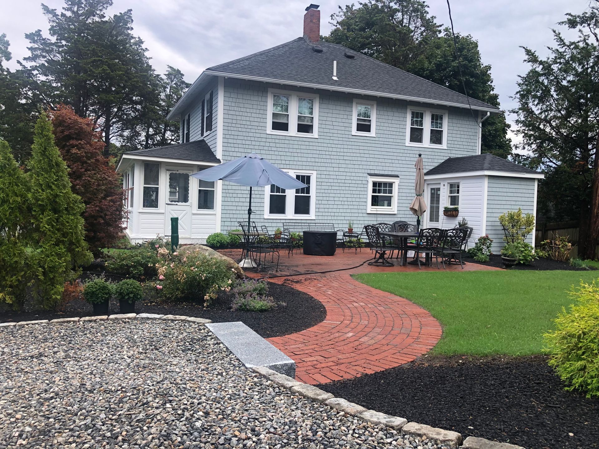 A gray house with a brick patio, outdoor seating, green lawn, and garden beds under a cloudy sky.