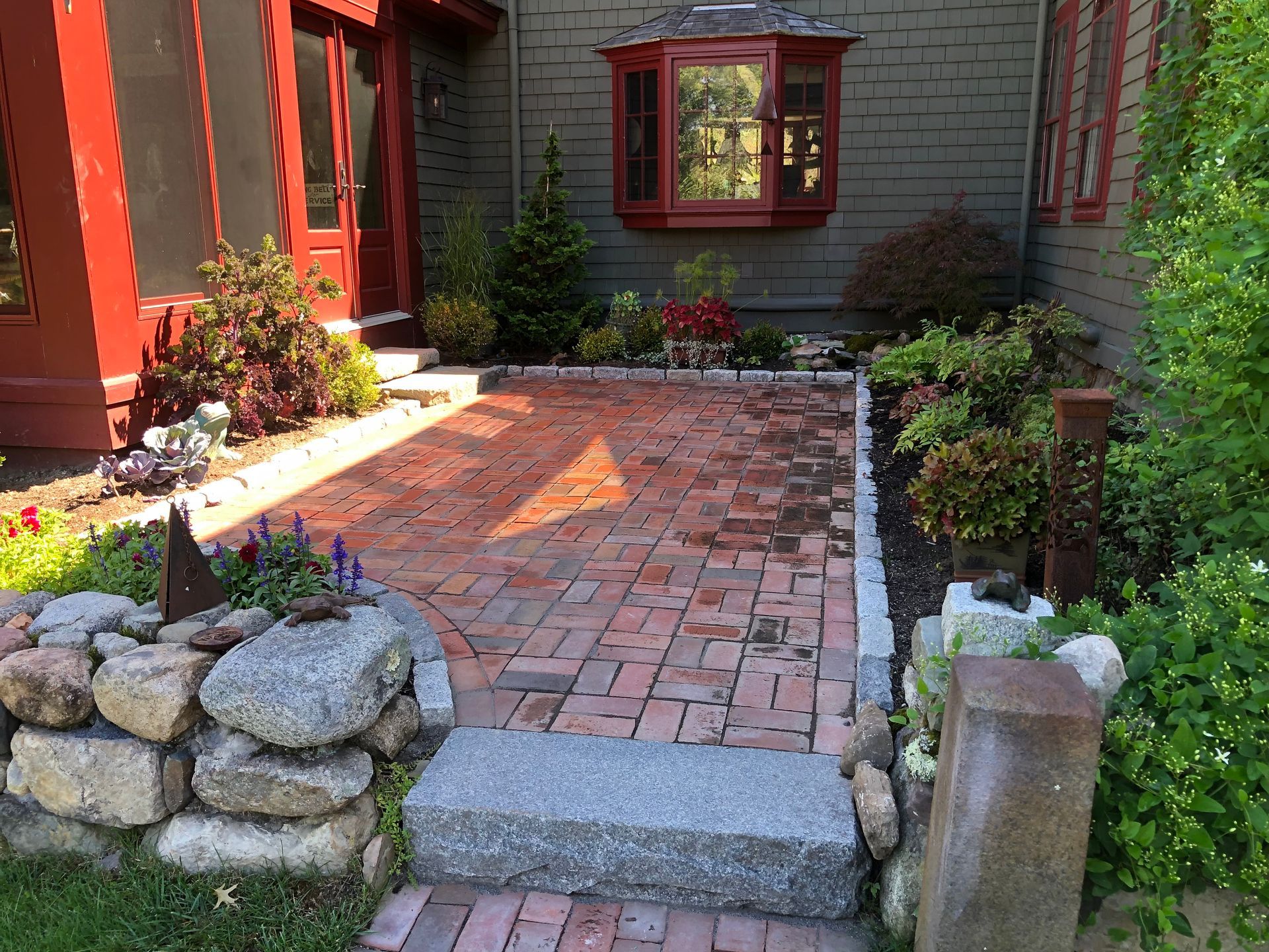 A brick patio framed by stone walls and garden greenery leads to a house with a red door and bay window.