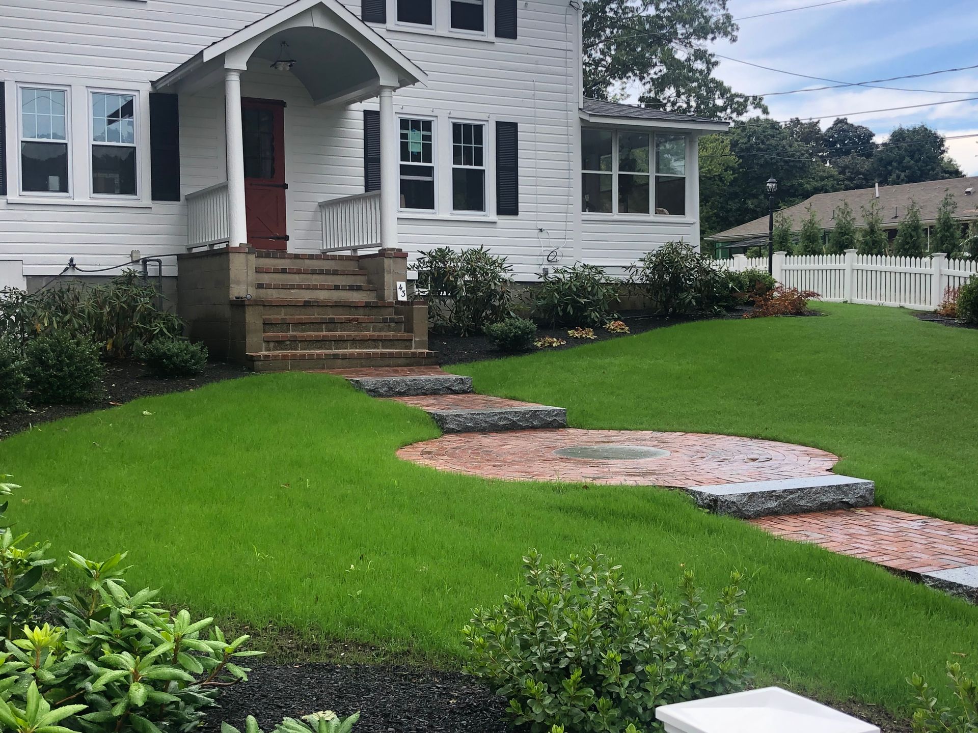 A white house with a brick front porch, steps, and a circular brick patio set within a green lawn.