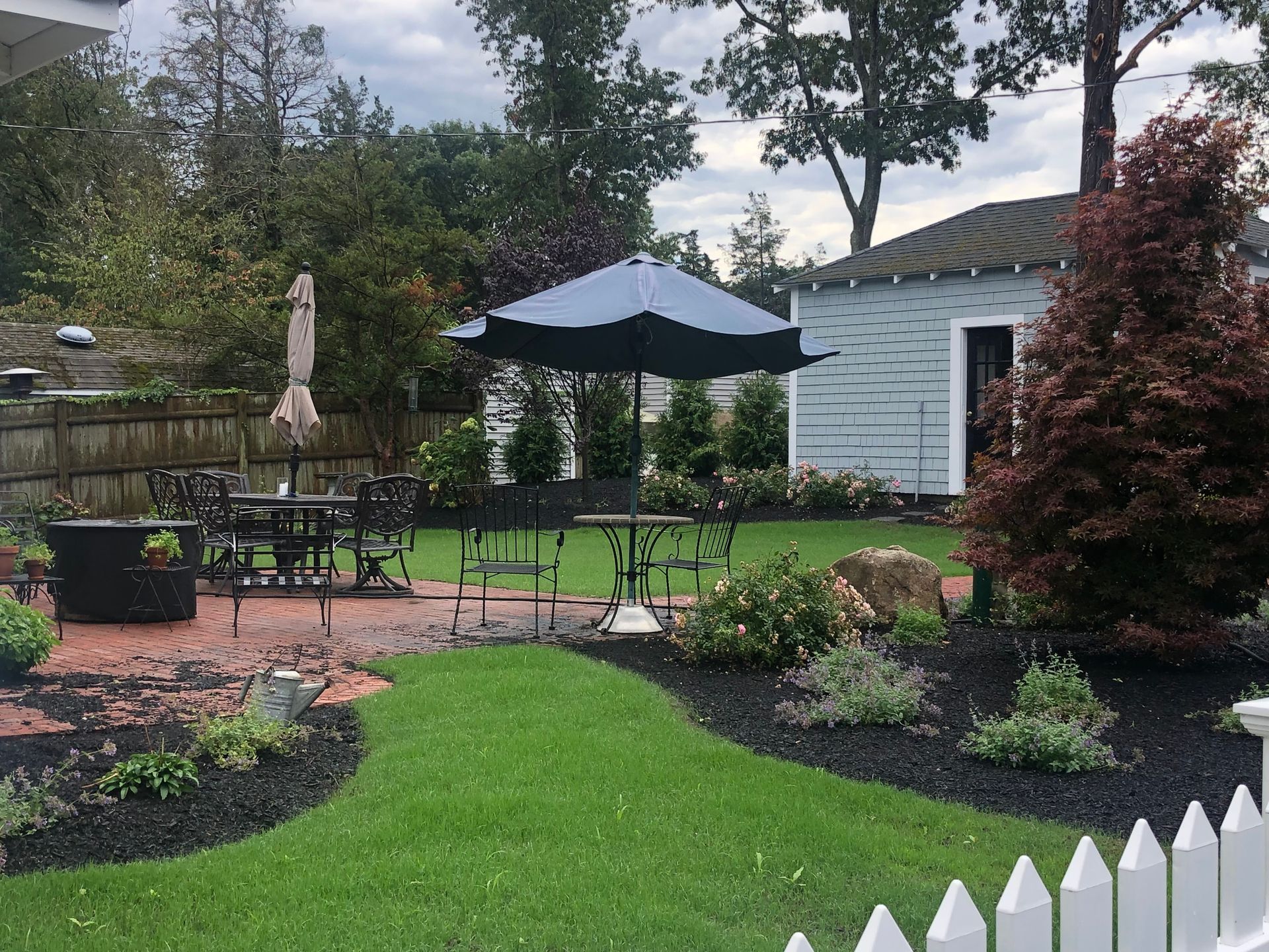A sunny backyard with a brick patio, outdoor dining set with an umbrella, green lawn, dark mulch beds, and a small shed.