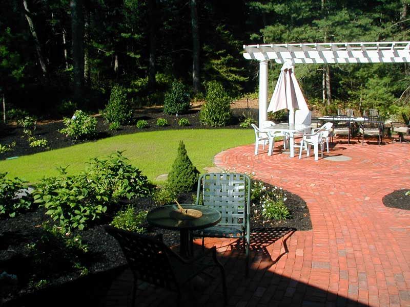 A brick patio with a white pergola, umbrella table, and chairs overlooking a green lawn bordered by dark mulched gardens.
