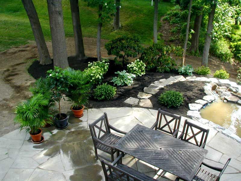 A patio with a wooden dining set, potted plants, and a small stone-lined pond in a wooded backyard.