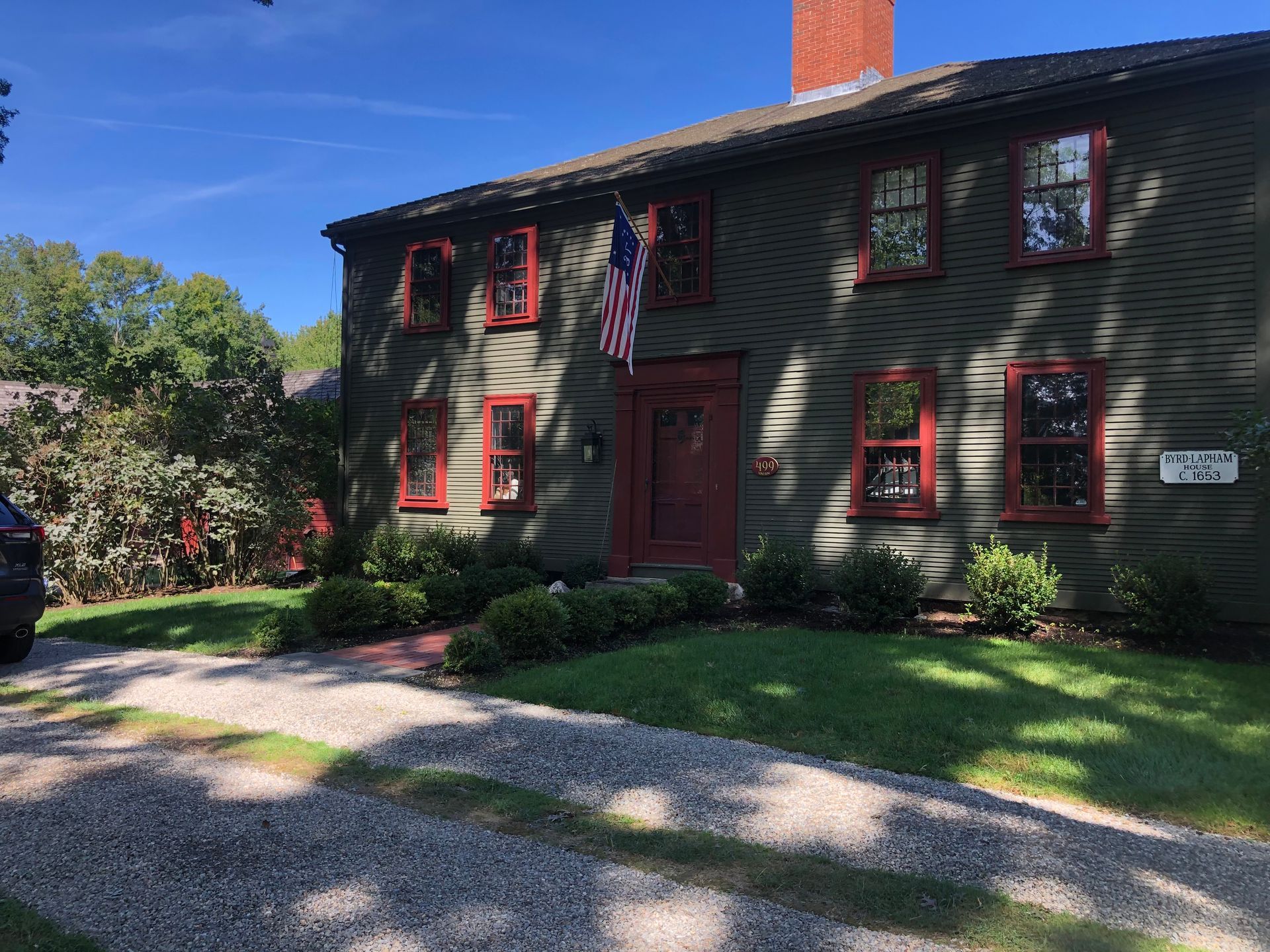 A two-story, grey colonial house with dark red window frames and trim, an American flag, and a gravel driveway.