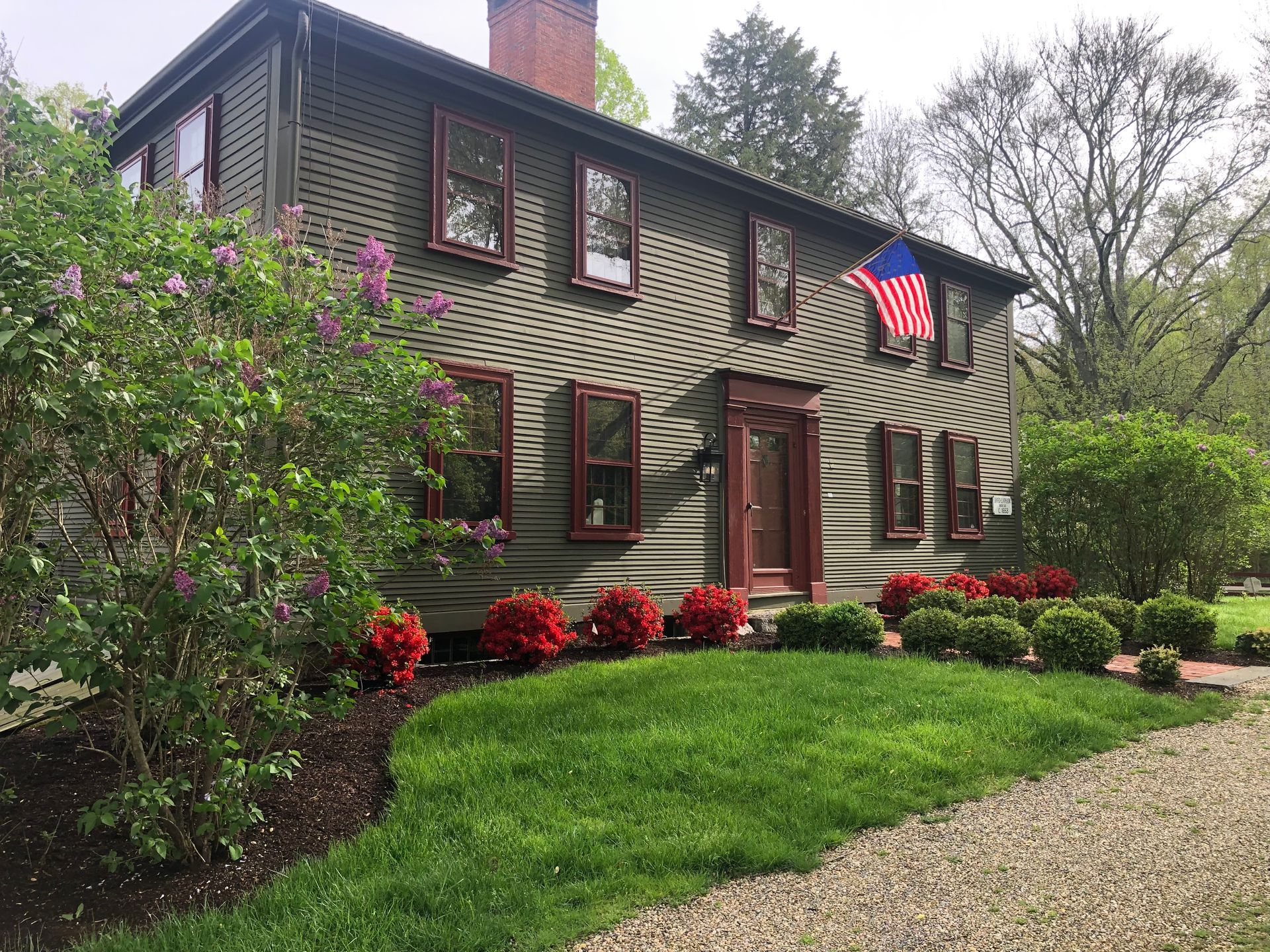 A historic two-story gray house with a red-trimmed door, dark shutters, and a front garden with a blooming lilac bush.