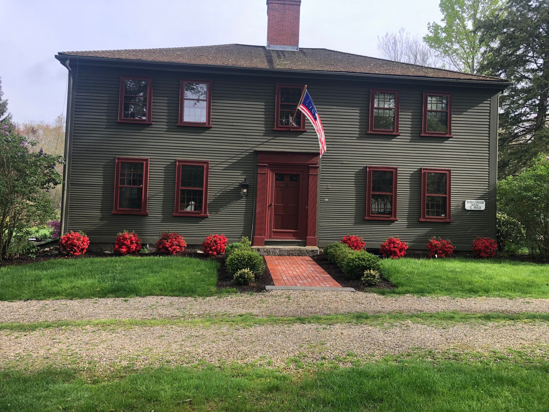 A two-story green historic home with a red door and window frames, a brick walkway, and a small American flag.