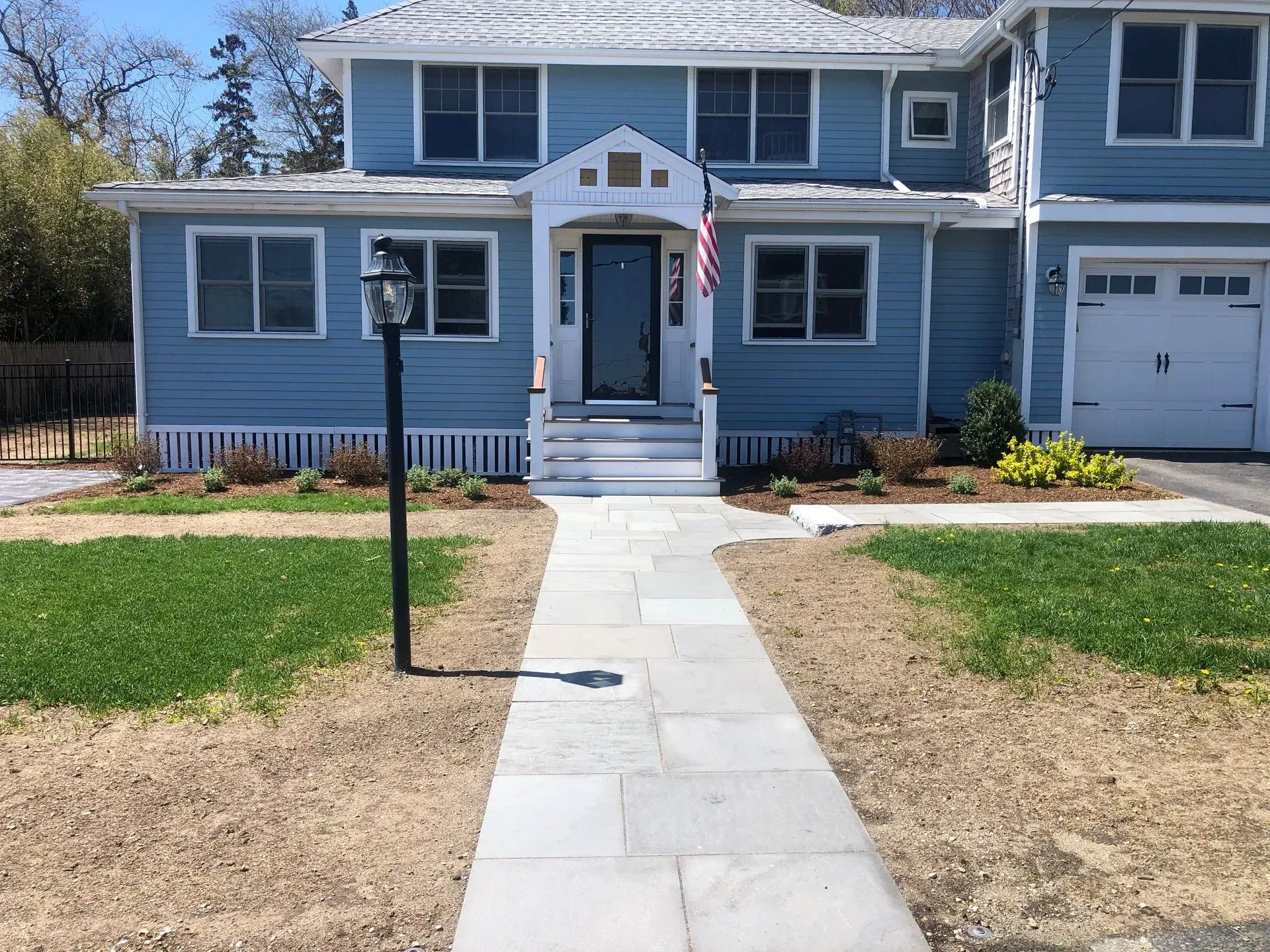 A blue, two-story house with a white front porch, a stone walkway, and a lamp post in a grassy front yard.