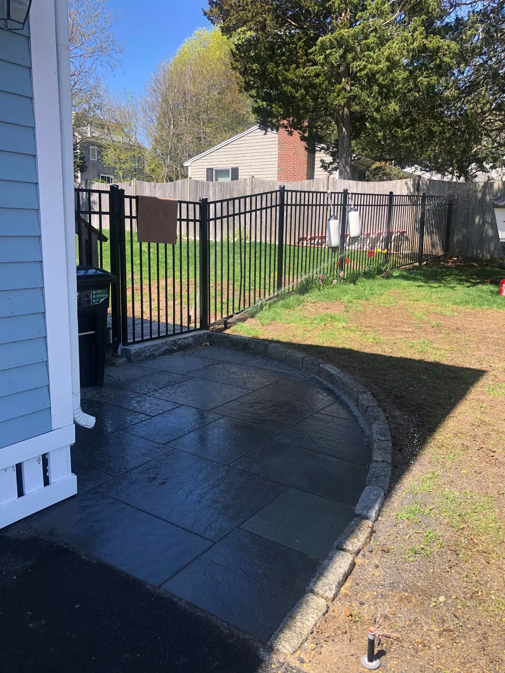 A curved stone patio outside a house with blue siding, bordered by a black metal fence and a grassy yard.