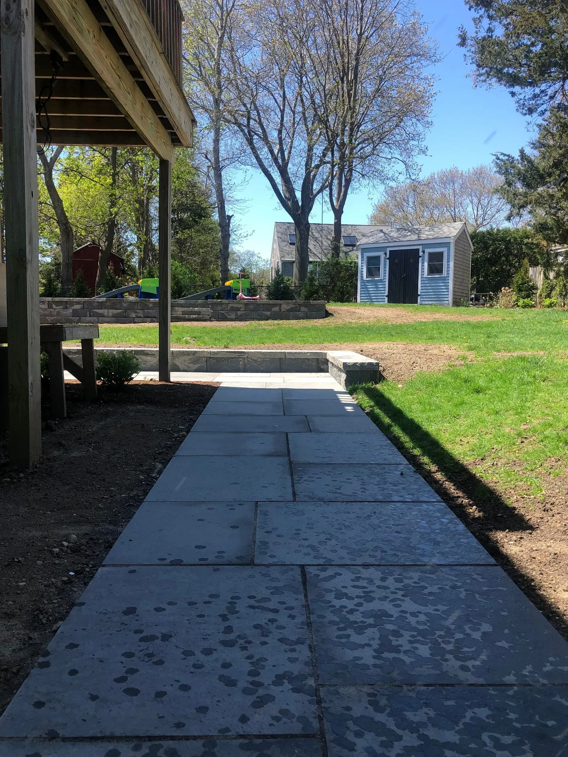 A stone walkway leads from a wooden deck toward a small shed in a sunny, green backyard.