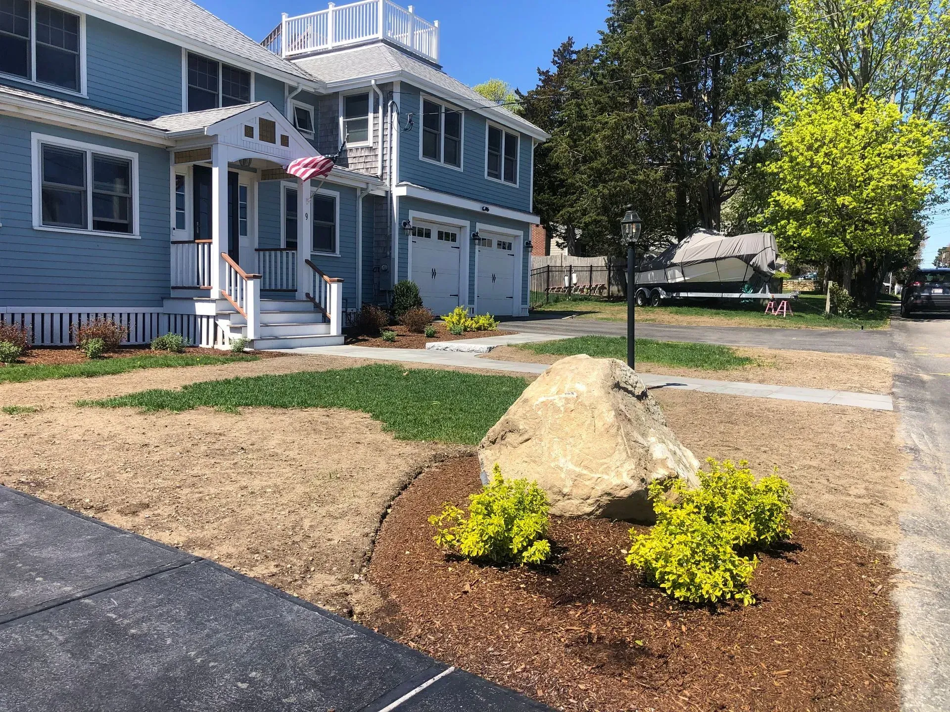 A blue two-story house with a white front porch and a landscaped front yard featuring a large rock and yellow bushes.