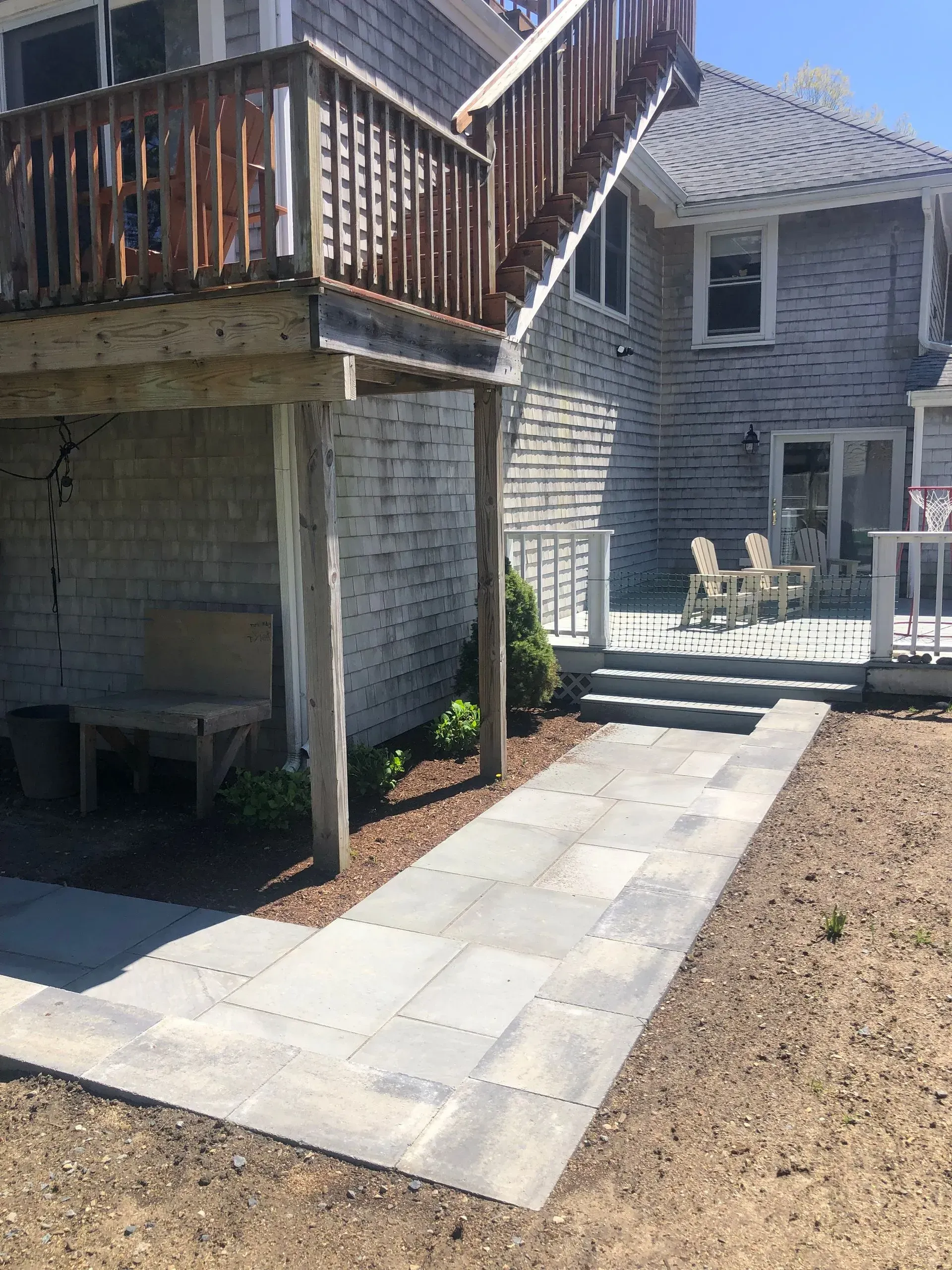 A light gray stone walkway leads to a backyard patio with chairs, set beneath a wooden deck and staircase.