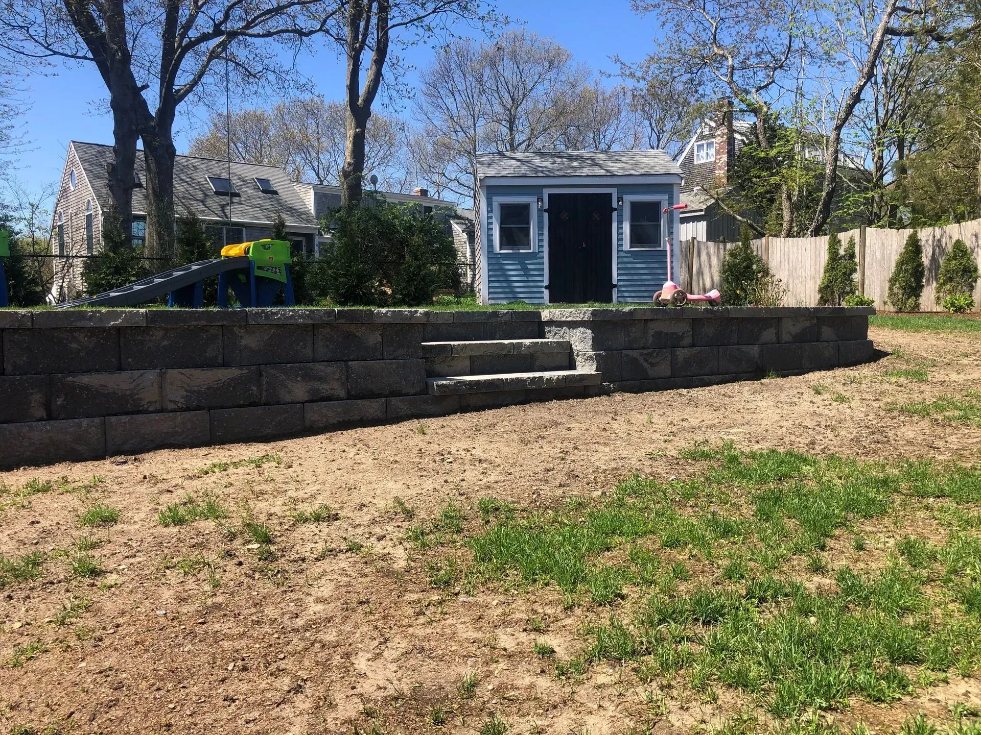 A stone retaining wall with integrated steps separates a sandy yard from a blue shed and nearby house under a clear sky.