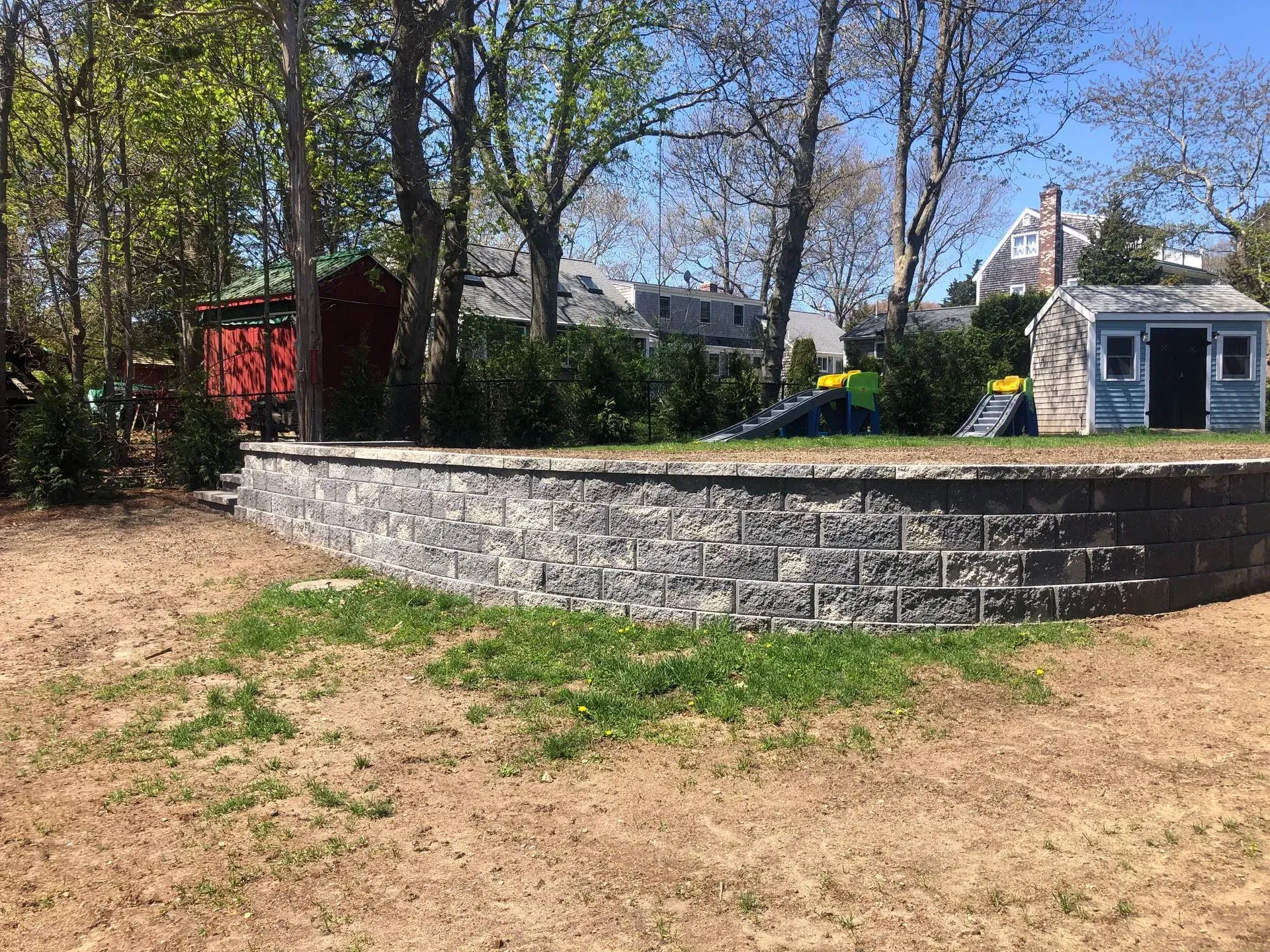 A stone retaining wall sits in a grassy yard, with a small blue shed, a red outbuilding, and trees in the background.