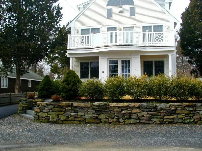 A two-story, cream-colored house with a stone retaining wall, balcony, and trimmed hedges in front.