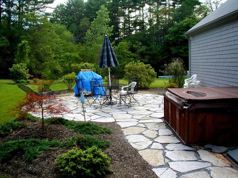 A stone patio with an umbrella-shaded table, a covered hot tub, and a landscaped garden next to a house.