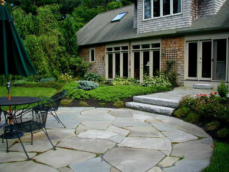 A stone patio with outdoor seating next to a shingled house with a landscaped garden.