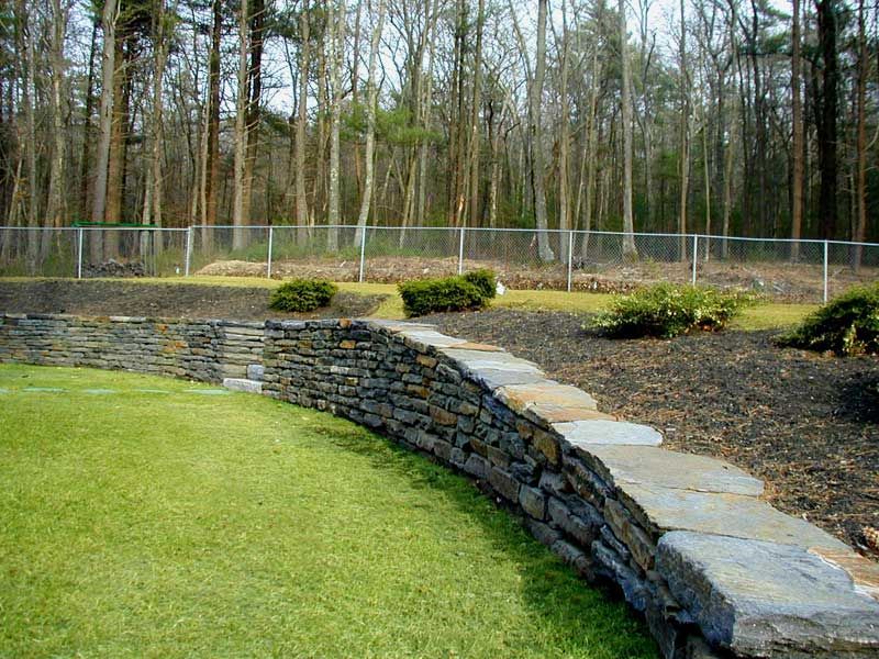 A curved stone retaining wall separates a green lawn from a garden bed with mulch and shrubs, backed by a fence and trees.
