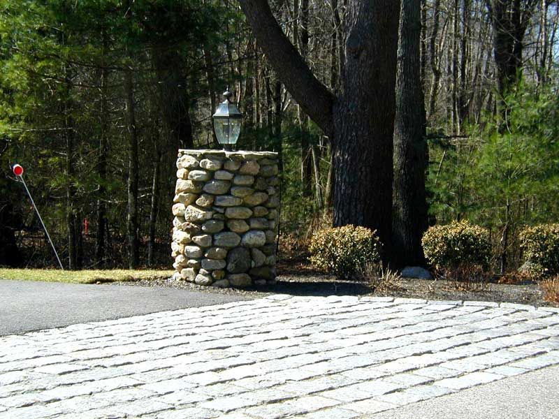 A cylindrical stone pillar with a lantern on top stands beside a patterned driveway in a wooded setting.