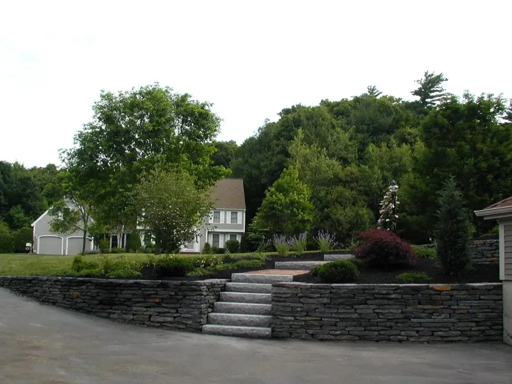 A stone retaining wall with a central staircase leads to a landscaped yard and a gray two-story house surrounded by trees.