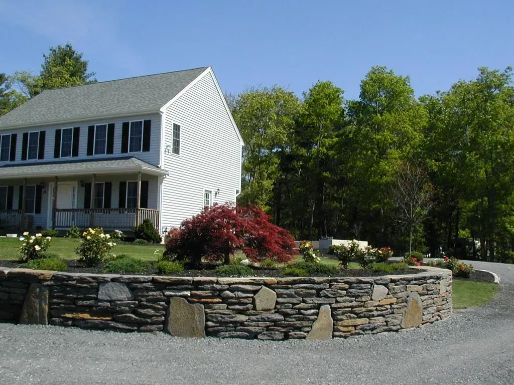 A two-story white house with a porch sits behind a decorative stone retaining wall and a red Japanese maple tree.