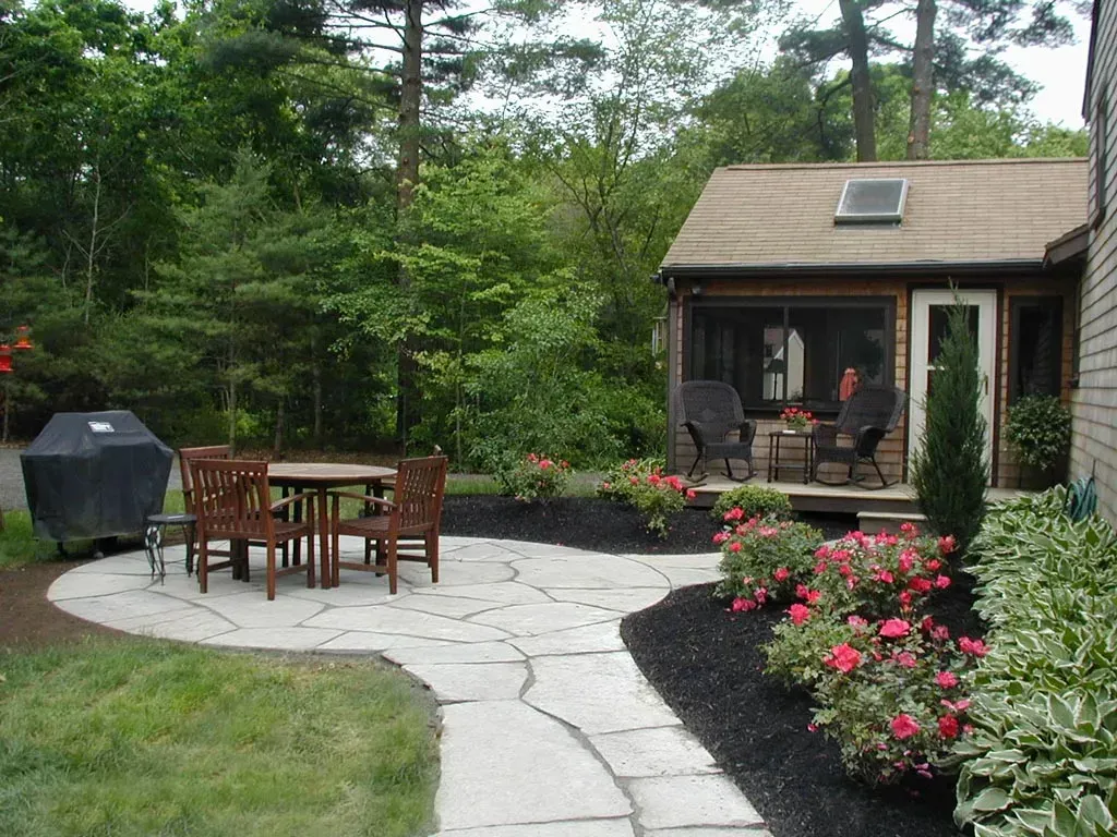A flagstone patio with a dining table and chairs, surrounded by garden beds, black mulch, and a small screened-in building.