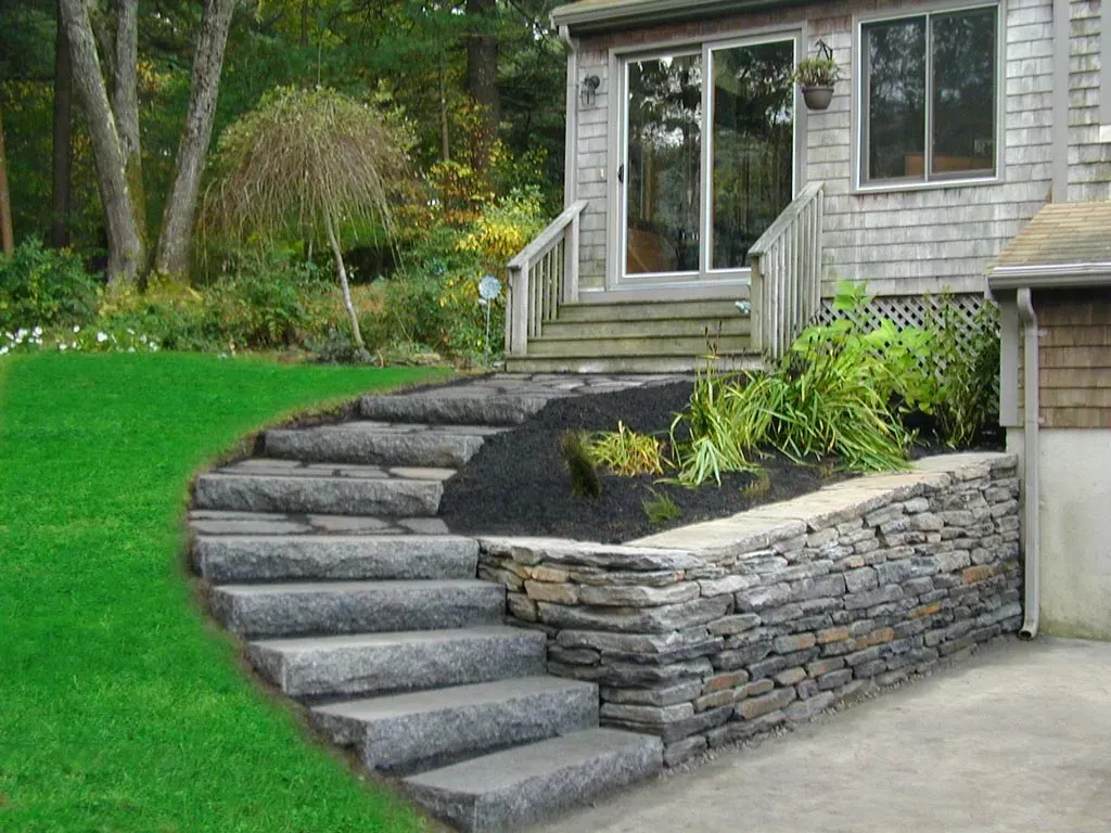 Stone steps curve from a grassy lawn up to a wooden deck and sliding glass door of a house with a stone retaining wall.