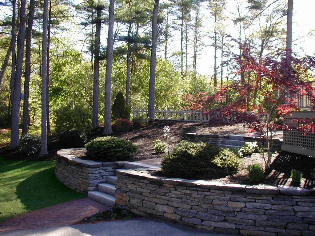 A stone retaining wall with steps leads to a mulched garden bed, manicured lawn, and trees under a sunny sky.