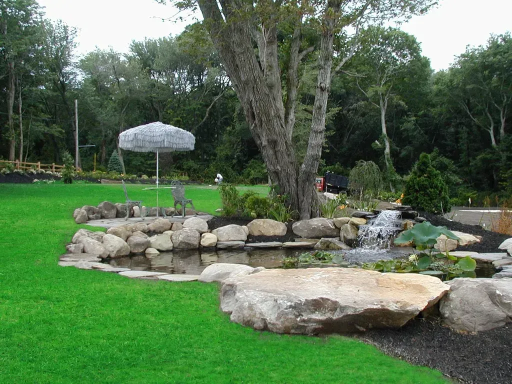A backyard stone pond with a small waterfall, surrounded by lush green grass, a large tree, and a patio with an umbrella.