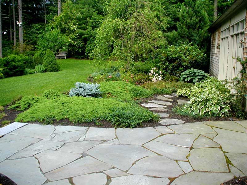 A flagstone patio leads into a lush, landscaped garden with green shrubs, perennials, and trees next to a garden shed.